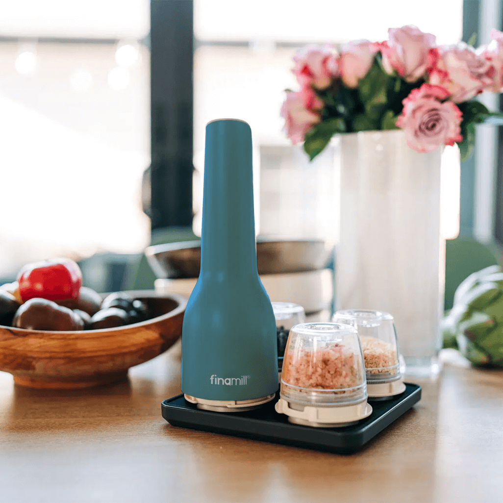 FINAMILL rechargeable grinder in Ocean Blue with spice containers on a kitchen table with flowers in the background.
