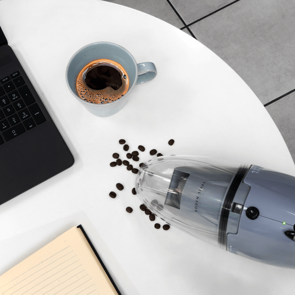 Boden Stark handheld vacuum cleaner on a table, cleaning coffee beans spilled near a laptop and a cup of coffee.