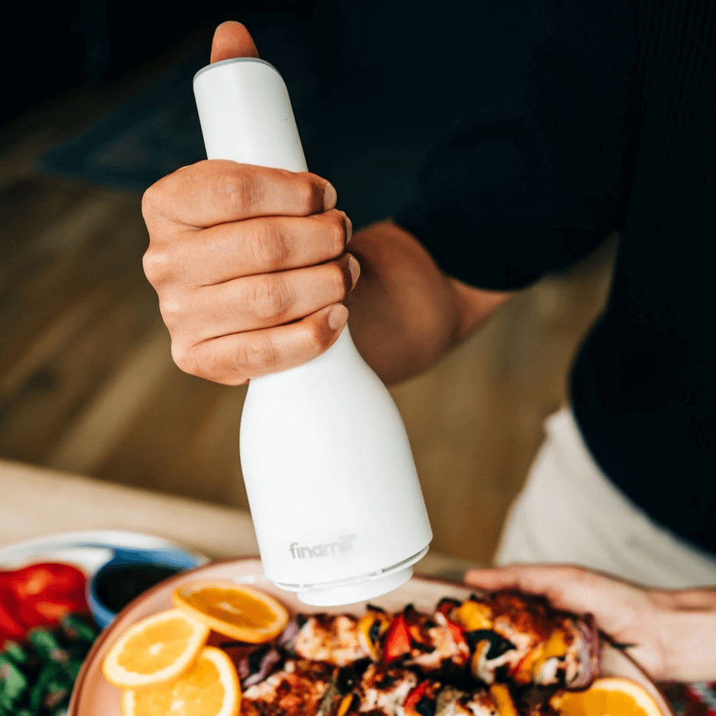 Hand holding FINAMILL rechargeable grinder in soft cream color over a plate of food.