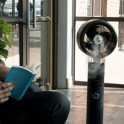 Person reading a book near the BODEN STARK 3-in-1 fan humidifier emitting mist in a sunny indoor space.