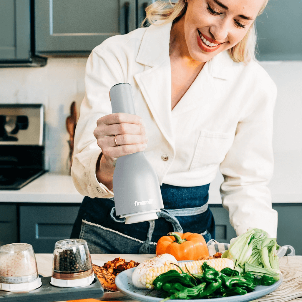Woman using FINAMILL RECHARGEABLE grinder - Stone in a modern kitchen with fresh vegetables on a table.
