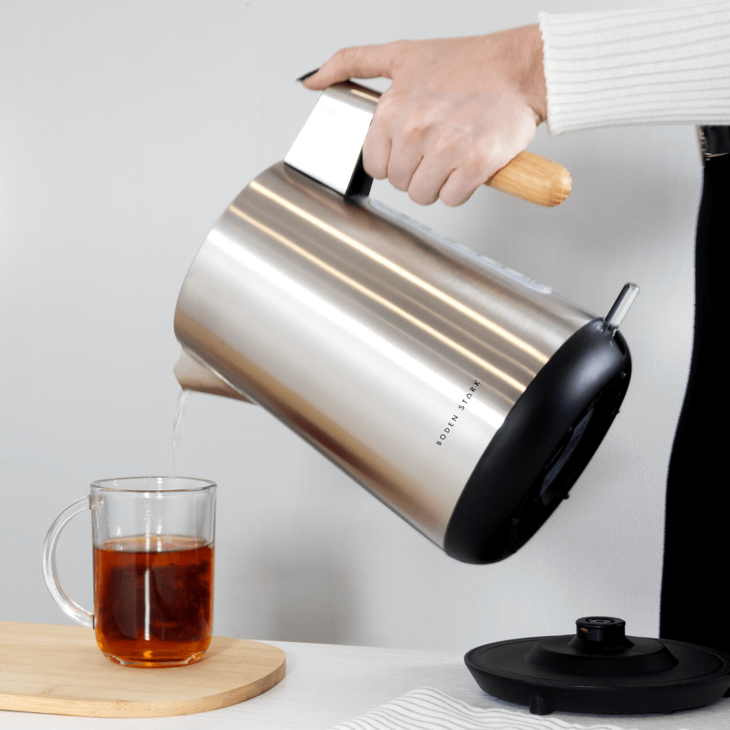 Boden Stark Anson Collection 1.7l stainless steel kettle pouring tea into a glass cup on a wooden cutting board.