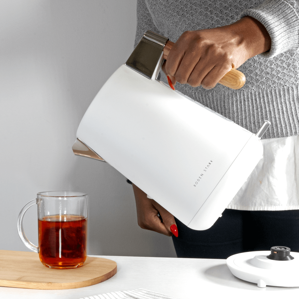 Boden Stark Anson Collection white kettle pouring tea into a glass cup on a wooden surface.